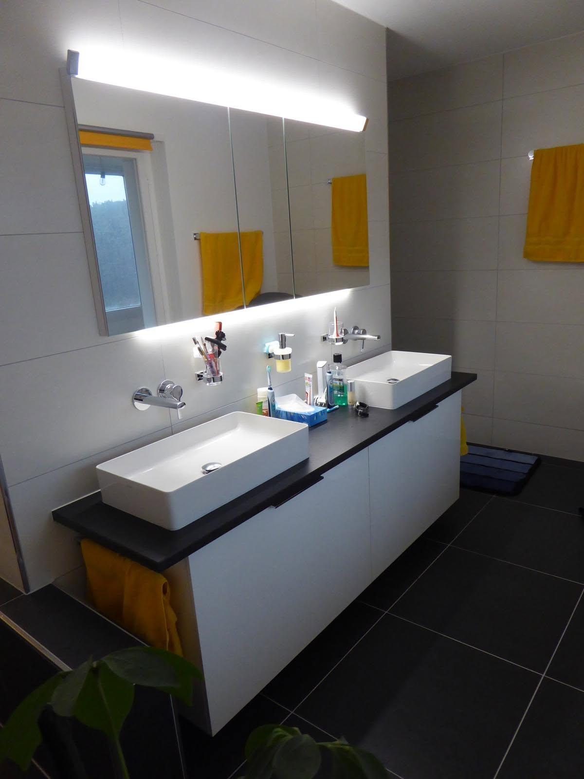 Badezimmer mit Doppelwaschbecken, beleuchtetem Spiegel und dunklen Bodenfliesen